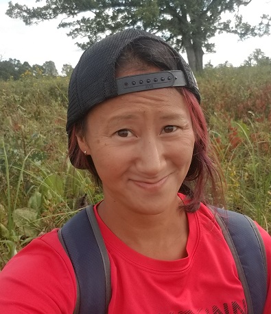 Nora Mitchell in a red shirt and black backwards baseball cap standing in field smiling at the camera