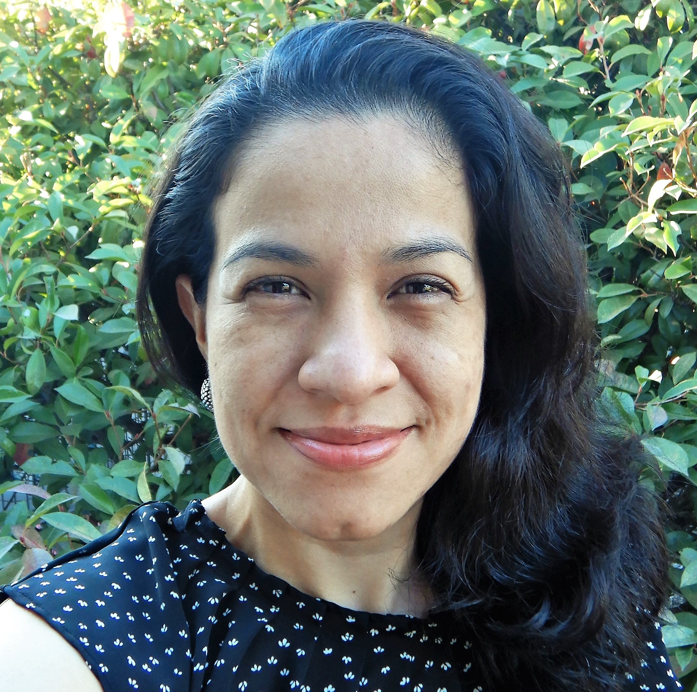 Headshot of Rosa Alicia Jiménez with long dark hair in front of a leafy green bush.